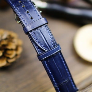 Puede incluir: Primer plano de una correa de reloj de cuero azul marino con textura de piel de cocodrilo. La correa tiene una hebilla y detalles de costura. El fondo est&aacute; borroso, con una pi&ntilde;a y una superficie de madera visibles.
