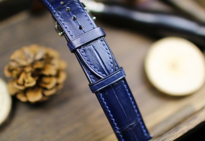 Puede incluir: Primer plano de una correa de reloj de cuero azul marino con textura de piel de cocodrilo. La correa tiene una hebilla y detalles de costura. El fondo est&aacute; borroso, con una pi&ntilde;a y una superficie de madera visibles.