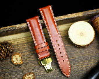 May include: Two brown leather watch straps with white stitching. The straps have a gold metal buckle and are laid out on a wooden surface.