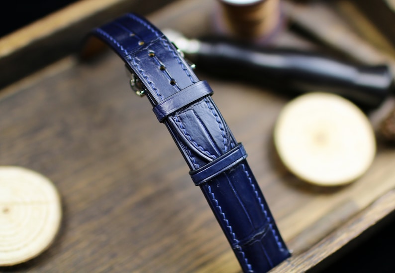 Puede incluir: Primer plano de una correa de reloj azul marino con un patr&oacute;n texturizado similar al de un cocodrilo. La correa tiene una hebilla y detalles de costura. La correa del reloj se muestra sobre una superficie de madera.