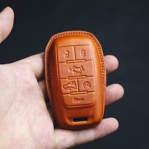 Peut inclure: Un porte-clés en cuir marron avec plusieurs boutons et symboles. Le porte-clés est conçu pour protéger une clé de voiture.