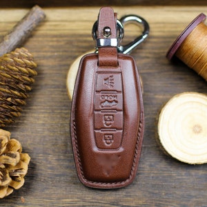 Może przedstawiać: Brązowy skórzany brelok do kluczy ze srebrnym metalowym kółkiem i karabińczykiem. Brelok ma cztery przyciski z symbolami do odblokowania, zablokowania, bagażnika i paniki.