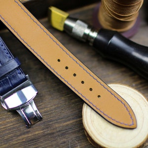 Puede incluir: Primer plano de una correa de reloj con interior de cuero beige y exterior azul marino. La correa est&aacute; cosida con hilo azul. Se ve una correa de reloj azul marino con un cierre plateado. Herramientas de artesan&iacute;a y una superficie de madera en el fondo.