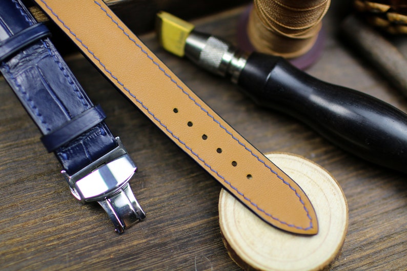 Puede incluir: Primer plano de una correa de reloj con interior de cuero beige y exterior azul marino. La correa est&aacute; cosida con hilo azul. Se ve una correa de reloj azul marino con un cierre plateado. Herramientas de artesan&iacute;a y una superficie de madera en el fondo.