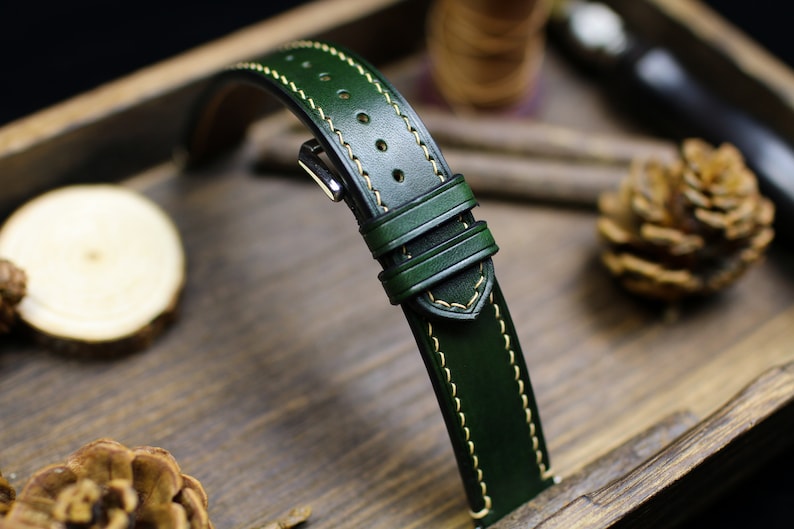 May include: A dark green leather watch strap with a light yellow stitching detail. The strap is shown on a wooden surface.
