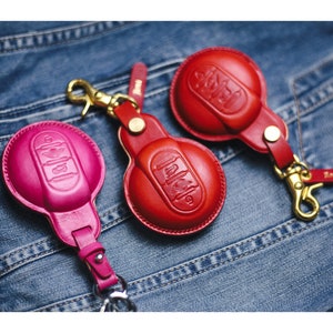 Op de afbeelding: Drie leren sleutelhangers in rood en roze, met een autosleutelontwerp. Elke sleutelhanger heeft een goudkleurige sluiting en een leren band. De sleutelhangers worden gepresenteerd op een denim achtergrond.