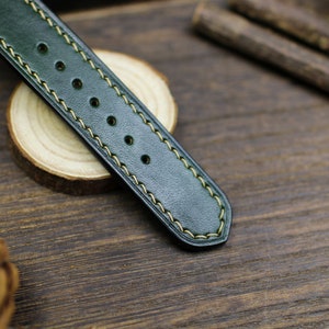May include: A dark green leather watch strap with yellow stitching. The strap has a smooth, textured surface and is designed for a wristwatch.