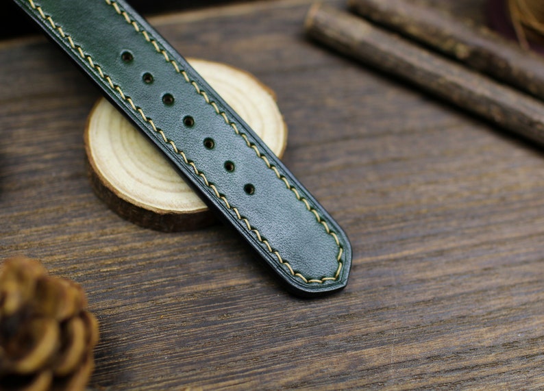 May include: A dark green leather watch strap with yellow stitching. The strap has a smooth, textured surface and is designed for a wristwatch.