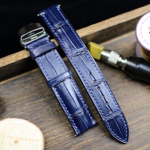 Puede incluir: Dos correas de reloj de cuero azul marino con efecto piel de cocodrilo. Las correas tienen costuras blancas y una hebilla plateada. Se presentan sobre una superficie de madera. Miden unos 20 cm.