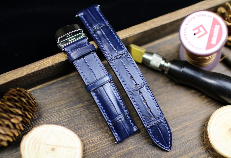 Puede incluir: Dos correas de reloj de cuero azul marino con efecto piel de cocodrilo. Las correas tienen costuras blancas y una hebilla plateada. Se presentan sobre una superficie de madera. Miden unos 20 cm.