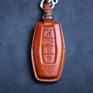 Op de afbeelding: Een bruine leren sleutelhanger met een zilveren sleutelring. De sleutelhanger heeft een gestikte rand en gegraveerde knoppen met de tekst "HOLD" en andere symbolen.