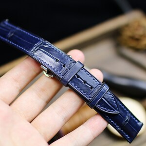 Puede incluir: Una correa de reloj azul marino con un patr&oacute;n de cocodrilo. La correa de cuero tiene costuras blancas y una hebilla plateada. La correa es una pieza de repuesto para un reloj de pulsera.