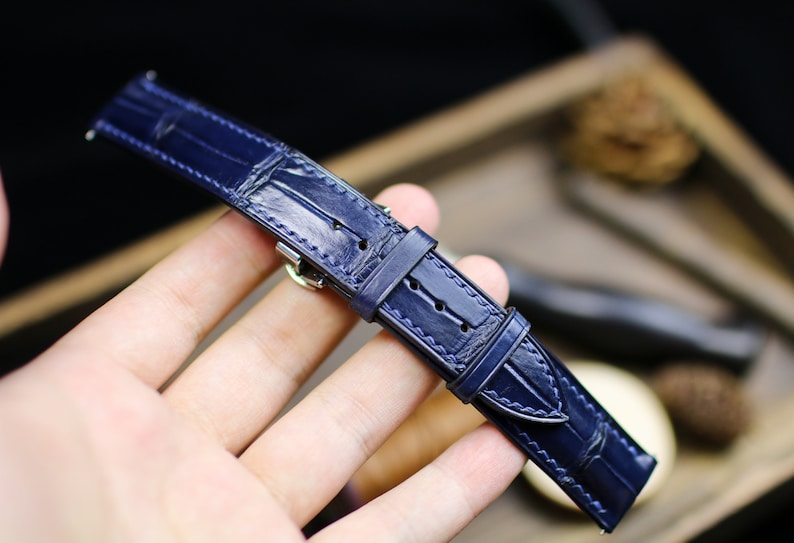 Puede incluir: Una correa de reloj azul marino con un patr&oacute;n de cocodrilo. La correa de cuero tiene costuras blancas y una hebilla plateada. La correa es una pieza de repuesto para un reloj de pulsera.