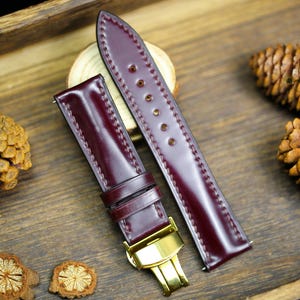 Op de afbeelding: Twee bordeauxrode leren horlogebandjes met gouden hardware. De bandjes hebben een gestikte rand en liggen op een houten oppervlak.