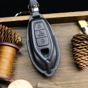 May include: Black leather car key fob cover with embossed buttons for lock, unlock, and trunk release. The key fob is on a wooden surface with a spool of thread, pine cones, and a leather crafting tool.