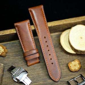 Op de afbeelding: Twee bruine leren horlogebandjes met oranje stiksels, een zilveren horlogesluiting en een zwarte horlogegesp worden gepresenteerd op een houten oppervlak. De bandjes zijn nieuw en ongebruikt. De achtergrond bevat houten schijfjes en decoratieve elementen.