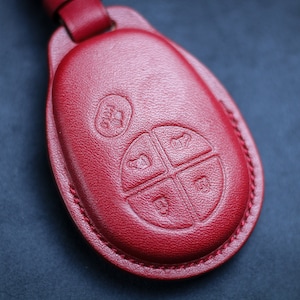 May include: Red leather key fob with four buttons and a small embossed logo. The fob has a stitched edge.