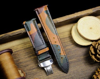 May include: Two camouflage-patterned leather watch straps. The straps feature a mix of green, brown, and orange hues. One strap is fastened with a silver clasp. The straps are displayed on a wooden surface.