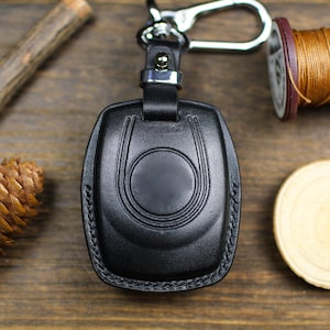 Può includere: Un portachiavi in ​​pelle nera con un anello argentato e un gancio argentato. Il portachiavi ha un bordo cucito e un design circolare al centro.