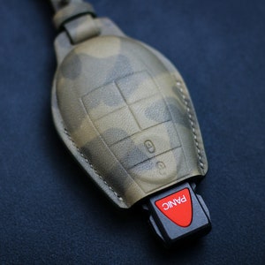 Pode incluir: Um chaveiro de couro com padrão de camuflagem com uma chave preta com um botão vermelho "PANIC".