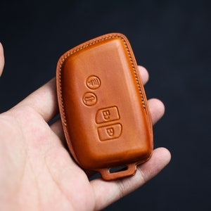 May include: A brown leather key fob with stitching around the edges. The fob has three buttons with symbols on them.