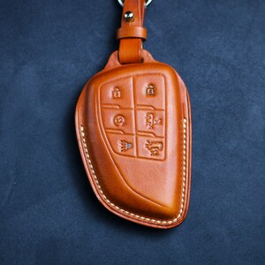 May include: A tan leather key fob cover with embossed icons for lock, unlock, boot, and other functions. The cover is stitched with white thread and attached to a leather strap and metal ring. The background is a dark blue.