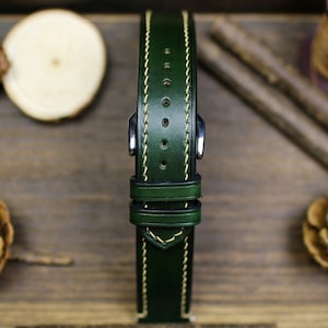 May include: A dark green leather watch strap with cream stitching. The strap has a buckle and two keeper loops.