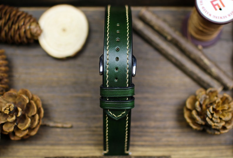 May include: A dark green leather watch strap with cream stitching. The strap has a buckle and two keeper loops.