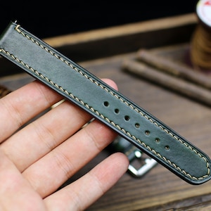 May include: A dark green leather watch strap with light beige stitching. The strap has five holes for adjusting the size.