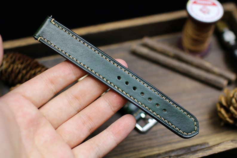 May include: A dark green leather watch strap with light beige stitching. The strap has five holes for adjusting the size.