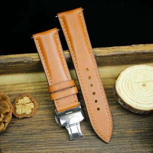 Puede incluir: Dos correas de reloj de cuero marrón con una hebilla plateada. Las correas están sobre una superficie de madera con una pequeña piña y un trozo de madera.