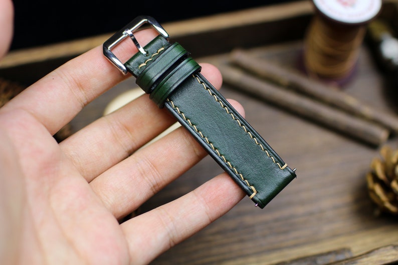 May include: A dark green leather watch strap with a silver buckle and yellow stitching.