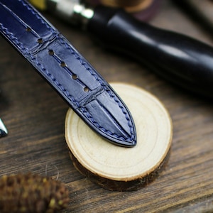 Puede incluir: Primer plano de una correa de reloj de cuero azul marino con un patr&oacute;n de cocodrilo y costuras azules. La correa est&aacute; sobre una superficie de madera con una rodaja de madera natural y herramientas de marroquiner&iacute;a en el fondo. Art&iacute;culo de lujo.