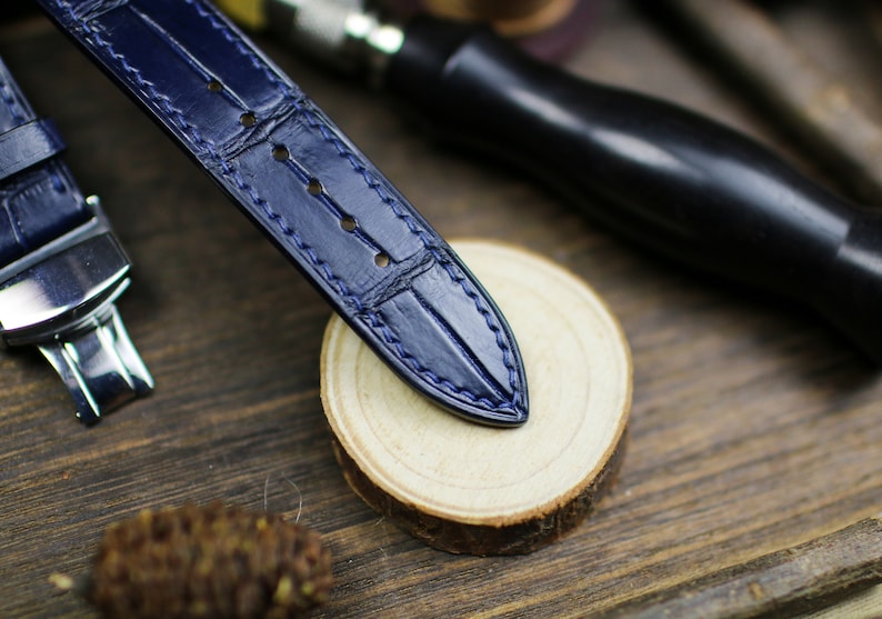 Puede incluir: Primer plano de una correa de reloj de cuero azul marino con un patr&oacute;n de cocodrilo y costuras azules. La correa est&aacute; sobre una superficie de madera con una rodaja de madera natural y herramientas de marroquiner&iacute;a en el fondo. Art&iacute;culo de lujo.