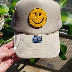 May include: A beige trucker hat with a yellow smiley face patch. The hat has a black mesh back and a black label that reads "OTTO WORLD FAMOUS FOAM FRONT OEM TRUCKER".