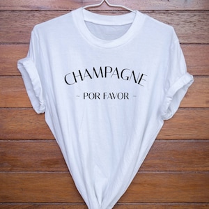Può includere: Maglietta bianca con testo nero che recita "Champagne - Por Favor -" su uno sfondo di legno.