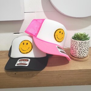 May include: Two trucker hats with a smiley face patch. One hat is white with a pink mesh back and the other is black with a white mesh back.