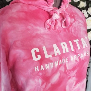 Puede incluir: Sudadera con capucha rosa tie-dye con un cord&oacute;n de ajuste blanco y el texto "CLARITA HANDMADE APPAREL" impreso en la parte delantera.