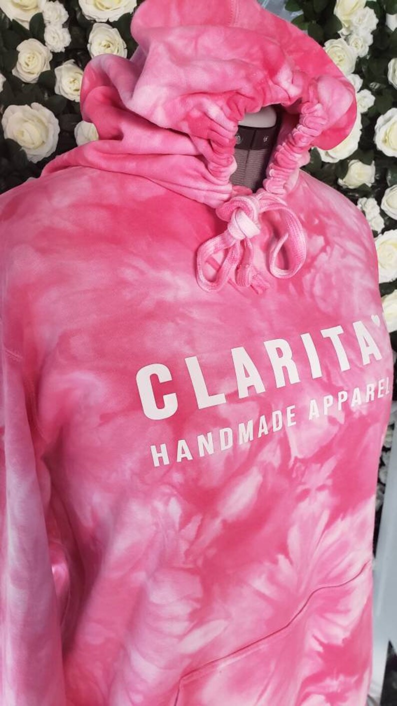 Puede incluir: Sudadera con capucha rosa tie-dye con un cord&oacute;n de ajuste blanco y el texto "CLARITA HANDMADE APPAREL" impreso en la parte delantera.