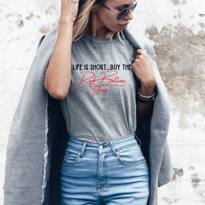 K&ouml;nnte beinhalten: Eine Frau tr&auml;gt ein graues T-Shirt mit dem Text "LIFE IS SHORT...BUY THE Red Bottom Shoes" in roten Buchstaben. Sie tr&auml;gt auch eine blaue Jeans und einen grauen Mantel.
