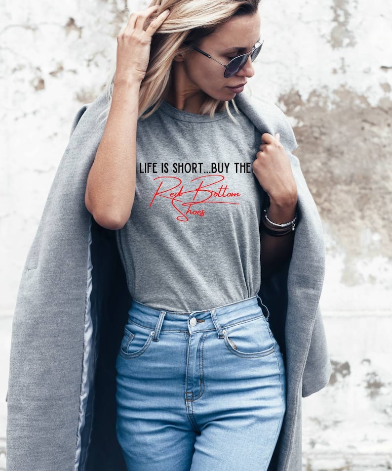 K&ouml;nnte beinhalten: Eine Frau tr&auml;gt ein graues T-Shirt mit dem Text "LIFE IS SHORT...BUY THE Red Bottom Shoes" in roten Buchstaben. Sie tr&auml;gt auch eine blaue Jeans und einen grauen Mantel.