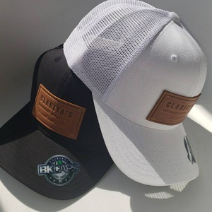 Pode incluir: Duas bonés de beisebol, um preto e um branco. Ambos os bonés têm um remendo de couro castanho com o texto "Clarita's Handmade Apparel Co.". O boné preto tem uma parte de trás de malha preta e um remendo preto com o texto "Original BK Caps Since 2003". O boné branco tem uma parte de trás de malha branca.