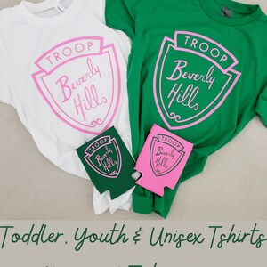 Puede incluir: Dos camisetas blancas y verdes con un dise&ntilde;o de escudo rosa que dice "TROOP Beverly Hills". Dos enfriadores de bebidas rosas y verdes a juego con el mismo dise&ntilde;o.