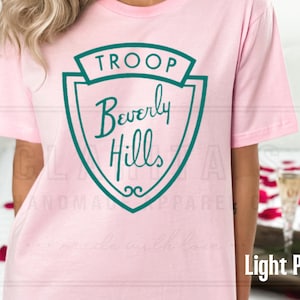 Puede incluir: Camiseta rosa claro con un gr&aacute;fico de escudo verde que dice "Troop Beverly Hills".