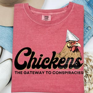 May include: A red t-shirt with a black graphic of a chicken wearing a tinfoil hat. The text "Chickens The Gateway to Conspiracies" is printed below the graphic.