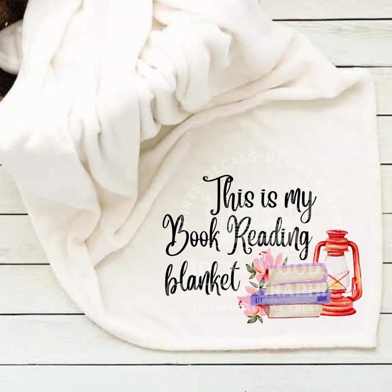 May include: White fleece blanket with the text "This is my Book Reading blanket" in black cursive font. A stack of books with a pink floral design and a red lantern are on the blanket.