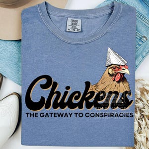 May include: A blue t-shirt with a black and white graphic of a chicken wearing a tinfoil hat. The text "Chickens The Gateway to Conspiracies" is printed below the image.