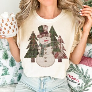 以下が含まれることがあります： クリーム色のTシャツ。チェック柄の帽子、スカーフ、ステッチのディテールが施された雪だるまのデザイン。雪だるまはチェック柄のクリスマスツリーの前に配置されています。「ROCK BLOOMERA」の文字が下にプリントされています。その人はジンジャーブレッドマンのマグカップを持っています。