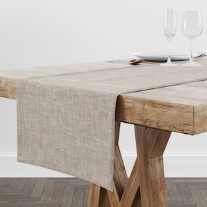 May include: A beige linen table runner on a wooden table. The runner is rectangular and has a textured appearance. Two wine glasses, a plate, and cutlery are set on the table, suggesting a dining setting.