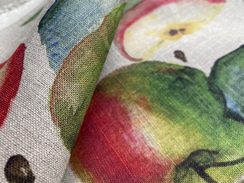 May include: Close-up of a fabric with a repeating pattern of red, green, and yellow apples. The fabric has a linen texture.